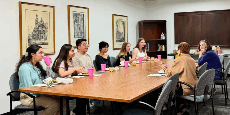 A group of students and faculty sit around a long conference table enjoying lunch and conversation with guest speaker Dr. Priscilla Meléndez in a casual setting.