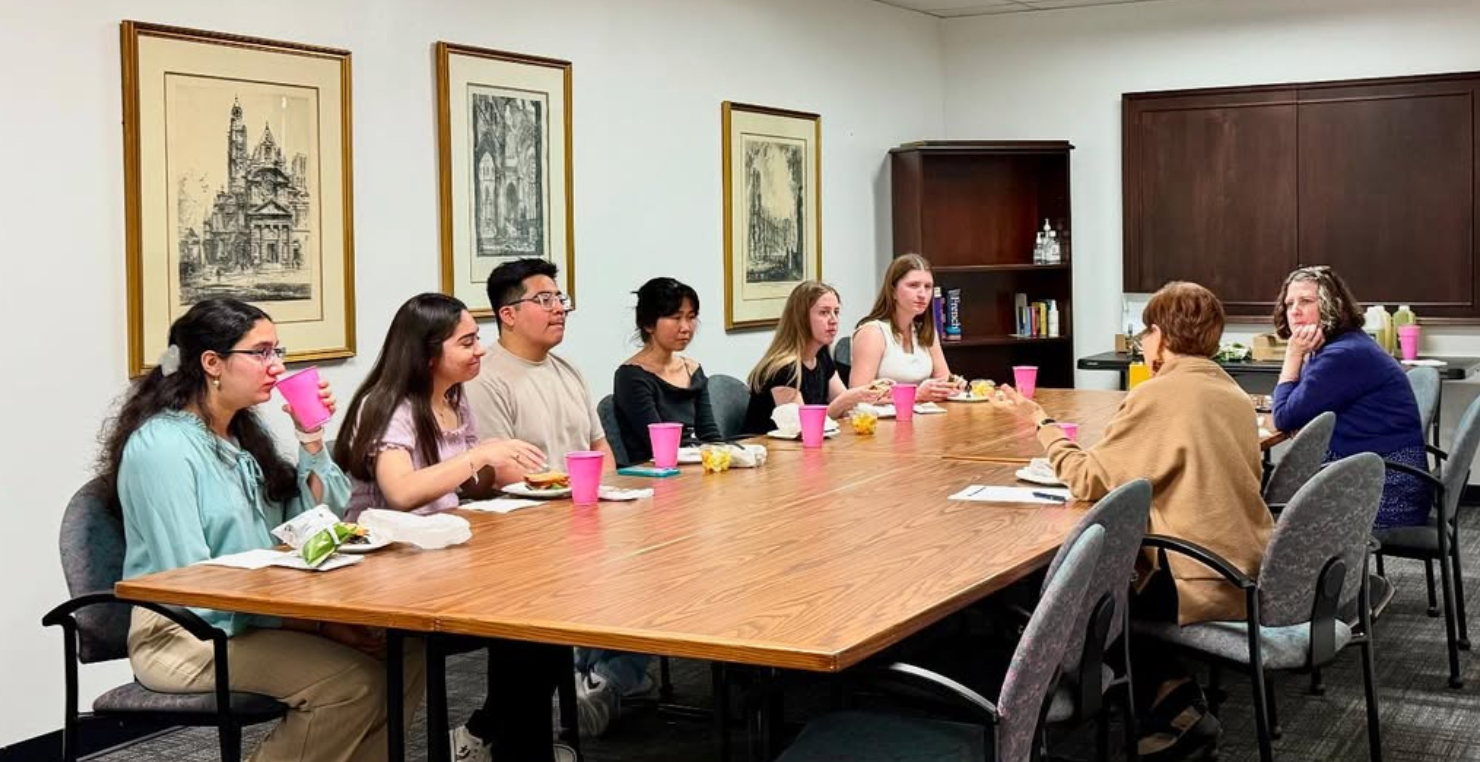 A group of students and faculty sit around a long conference table enjoying lunch and conversation with guest speaker Dr. Priscilla Meléndez in a casual setting.