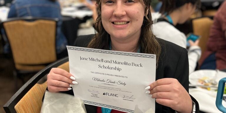 Alt Text: A young woman with long, wet-look hair smiles while holding a certificate for the Jane Mitchell and Manolita Buck Scholarship. She is wearing a black blazer and a red tie. In front of her is a bouquet of flowers wrapped in white paper. The background shows a conference setting with people sitting at tables engaged in discussions. A smartphone with a floral case rests on the table next to her.