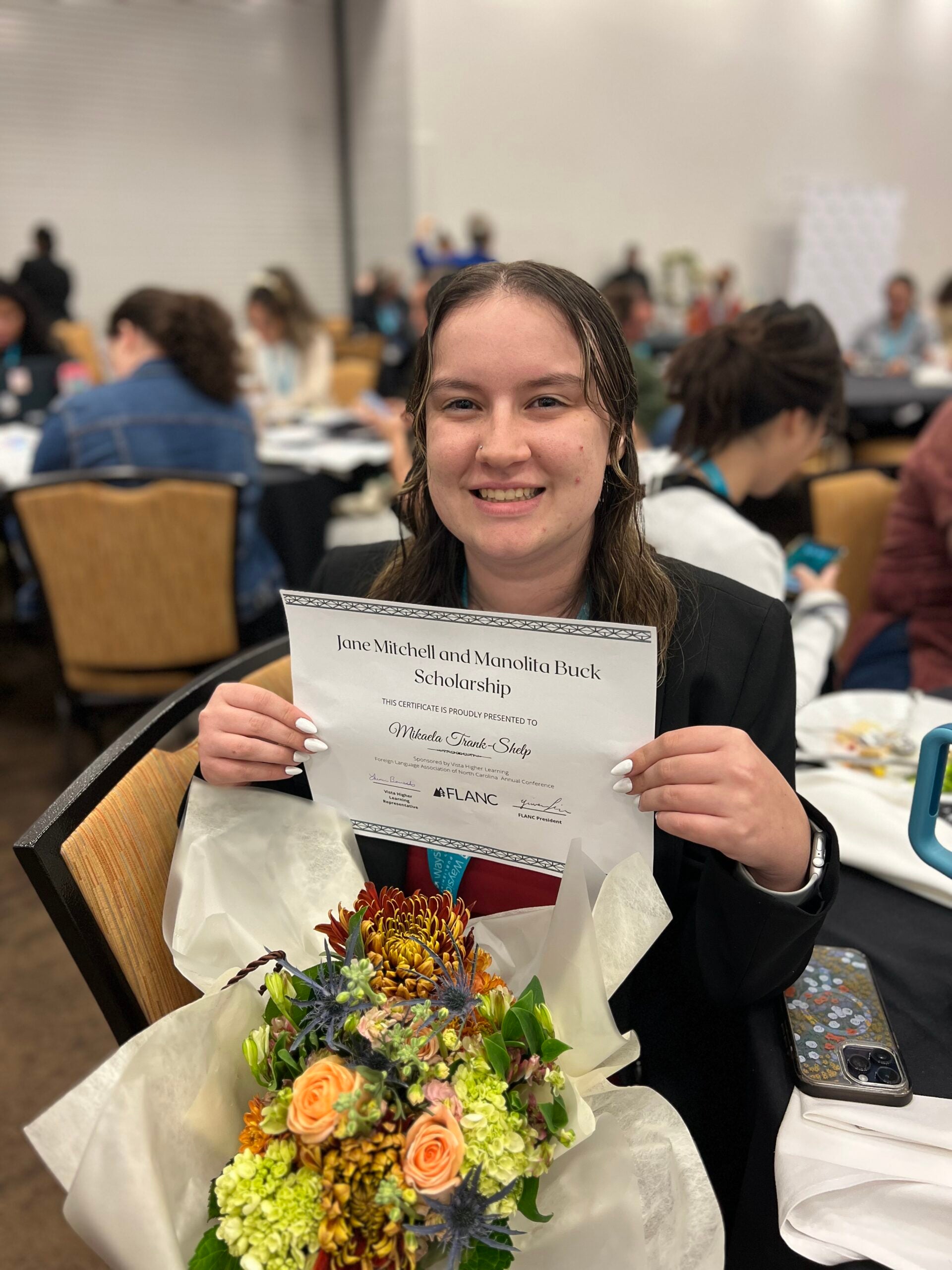 Alt Text: A young woman with long, wet-look hair smiles while holding a certificate for the Jane Mitchell and Manolita Buck Scholarship. She is wearing a black blazer and a red tie. In front of her is a bouquet of flowers wrapped in white paper. The background shows a conference setting with people sitting at tables engaged in discussions. A smartphone with a floral case rests on the table next to her.