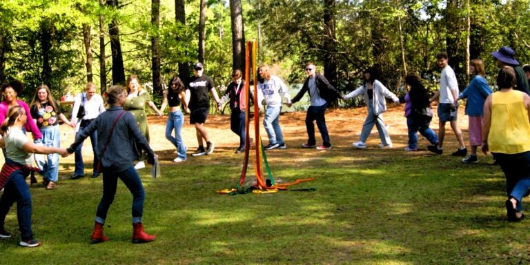 A group of students and community members participate in a traditional Maypole dance at River Park North, holding colorful ribbons as they walk around a central pole under the trees.
